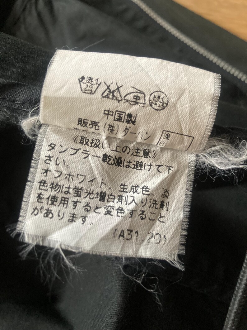 May include: A white rectangular garment care label sewn onto black fabric. The label features black Japanese text, including instructions to avoid tumble drying and warnings about color changes with fluorescent detergents, along with laundry symbols.