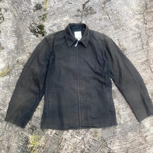 May include: A charcoal gray zip-up jacket featuring a classic collar, long sleeves, and a central metal zipper closure. The garment has a structured fit with visible vertical seam detailing along the front panels and a small white label at the neck.
