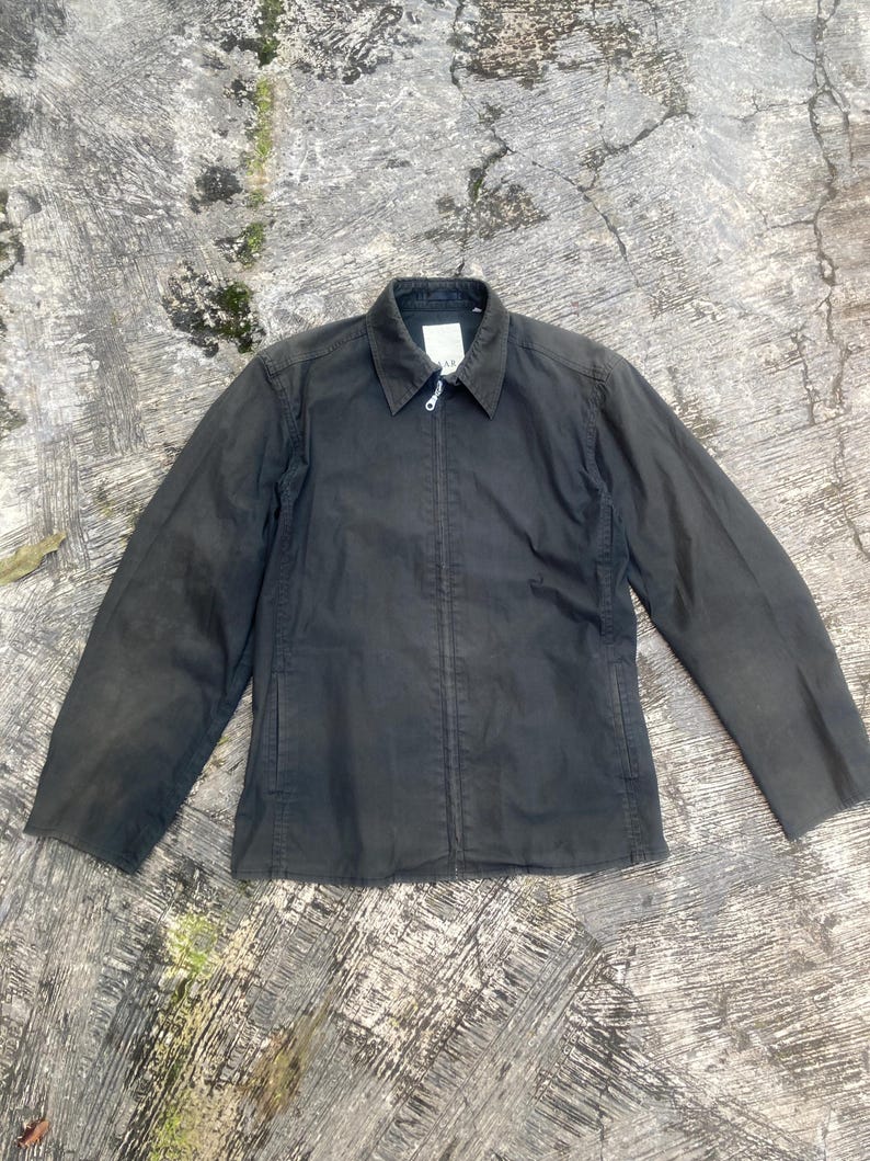 May include: A charcoal gray zip-up jacket featuring a classic collar, long sleeves, and a central metal zipper closure. The garment has a structured fit with visible vertical seam detailing along the front panels and a small white label at the neck.