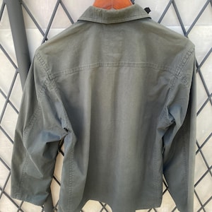 May include: A long-sleeved olive green button-down shirt featuring a classic collar, a horizontal back yoke seam, and visible stitching along the sleeves and sides. The shirt is displayed on a wooden hanger against a dark metal lattice structure.