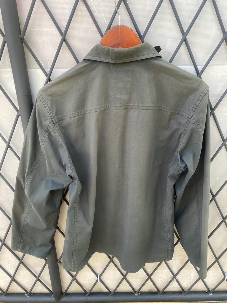 May include: A long-sleeved olive green button-down shirt featuring a classic collar, a horizontal back yoke seam, and visible stitching along the sleeves and sides. The shirt is displayed on a wooden hanger against a dark metal lattice structure.