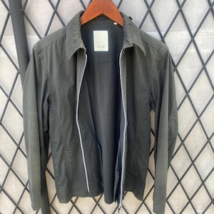 May include: A dark olive green jacket with a full-length silver zipper, a standard collar, and long sleeves. The interior label features the text A.A.R. The jacket is constructed with a structured fabric and includes two vertical side pockets.