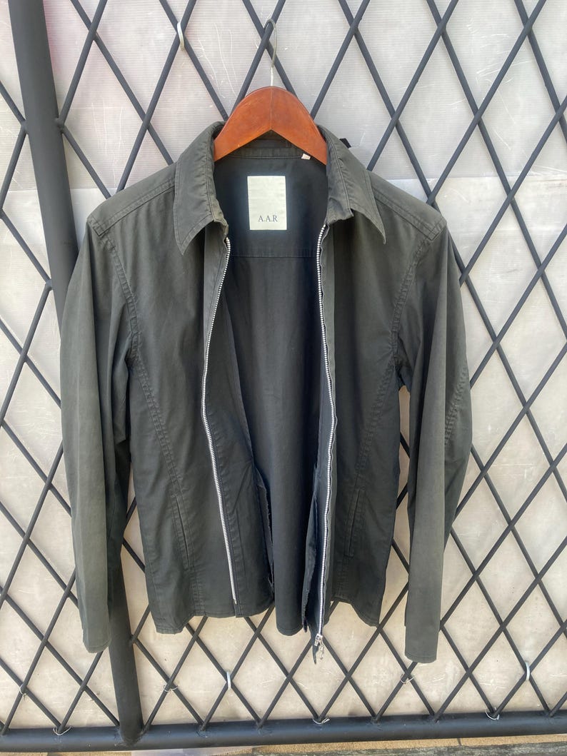 May include: A dark olive green jacket with a full-length silver zipper, a standard collar, and long sleeves. The interior label features the text A.A.R. The jacket is constructed with a structured fabric and includes two vertical side pockets.