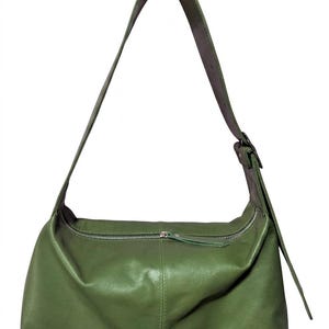 May include: Olive green leather shoulder bag with a long adjustable strap. The bag features a silver zipper closure and a simple, minimalist design. The bag is approximately 12 inches wide.