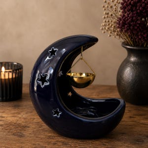 Puede incluir: Un quemador de aceite de cerámica azul oscuro con forma de media luna y recortes en forma de estrella. Un pequeño cuenco de metal dorado brillante cuelga de la parte superior de la luna mediante un fino soporte, sobre una base para una vela.