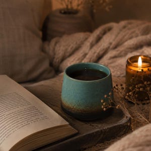 Peut inclure: Une tasse en céramique avec une finition dégradée passant d'un bleu sarcelle clair au niveau du bord à une base brun foncé. La tasse est remplie d'un liquide sombre et repose sur un plateau en bois foncé à côté d'un livre ouvert et d'une bougie.