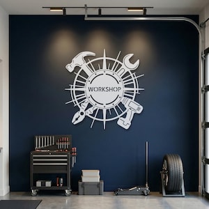 May include: A white metal wall sign featuring a circular center with the word WORKSHOP cut out, surrounded by a radial design of tools including a hammer, wrench, pliers, and a cordless drill, mounted on a dark blue wall in a garage setting.