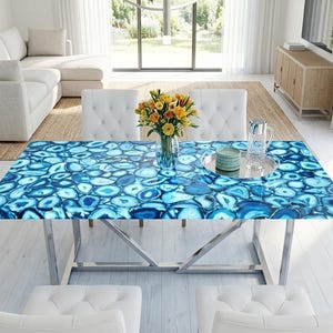 Puede incluir: Una mesa de comedor rectangular con un tablero compuesto por numerosas rodajas de ágata azul con acabado pulido, sostenida por una estructura de metal plateado con patas anguladas, en una habitación luminosa con suelo y muebles blancos.