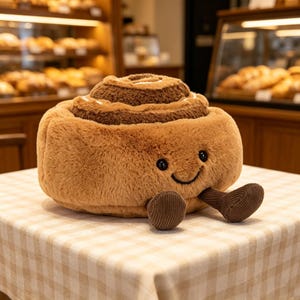 Puede incluir: Un peluche de rollo de canela marrón con una cara sonriente y pies de pana. El juguete suave tiene un diseño en espiral y está sobre un mantel a cuadros. El fondo muestra una panadería con pan.