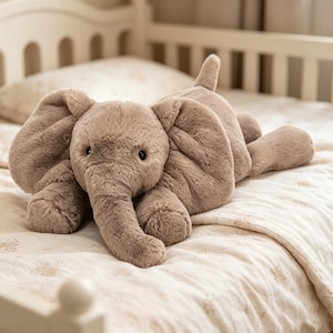 Op de afbeelding: Een zacht, taupekleurig olifantenspeelgoed met grote, slappe oren, een slurf en kleine zwarte ogen. Het knuffeldier ligt op een bed met een patroon dekbed en een witte hoofdbord op de achtergrond.