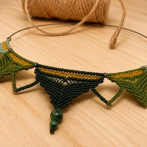 Op de afbeelding: Een macramé ketting met een reeks driehoekige geweven segmenten in tinten donkergroen, olijf en licht saliegroen, geaccentueerd met mosterdgeel garen. Het centrale deel bevat twee donkergroene gespikkelde kralen die naar beneden hangen.