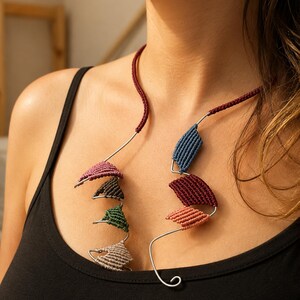 Puede incluir: Un collar con un cordón rojo oscuro y una estructura de alambre plateado. El alambre está adornado con varios elementos pequeños de macramé en forma de rombo de diversos colores, incluyendo rosa empolvado, marrón oscuro, verde, beige, azul y granate.