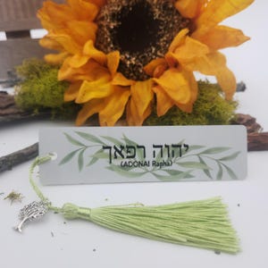 May include: A rectangular bookmark with a light gray background featuring green leaf illustrations and the Hebrew text "יהוה רפא" with "(ADONAI RAPHA)" printed below. It includes a light green thread tassel and a silver tree charm.