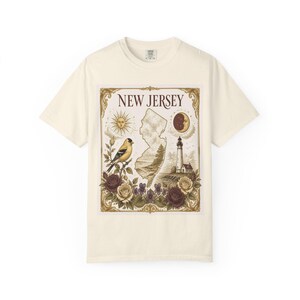May include: A cream-colored t-shirt featuring a rectangular graphic with the text NEW JERSEY. The design includes a map of New Jersey, a sun, a moon, a lighthouse, a goldfinch on a branch, and a collection of red, white, and purple roses.