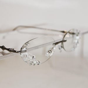 Può includere: Occhiali ovali senza montatura con aste in metallo color argento e lenti trasparenti decorate con piccoli elementi sfaccettati simili a cristalli lungo i bordi esterni. Un sottile ponte in metallo collega le lenti, dotate di naselli regolabili.
