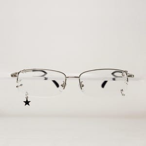 Könnte beinhalten: Silberfarbene Halbrandbrille mit klaren Gläsern und einem dünnen Metallrahmen. Ein kleiner, hängender schwarzer Sternanhänger ist an der unteren äußeren Ecke des linken Glases befestigt, während das rechte Glas einen kleinen silbernen Sternanhänger aufweist.
