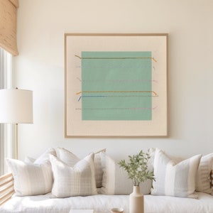 May include: A light wood daybed with white linen cushions and a light wood coffee table with a tray and a stack of books. The daybed is in front of a window with a view of the ocean and a green abstract painting hangs on the wall.