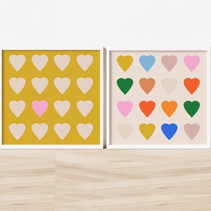 Op de afbeelding: Twee ingelijste prints met een patroon van harten. De print aan de linkerkant heeft een gele achtergrond met beige harten, één roze hart in het midden. De print aan de rechterkant heeft een beige achtergrond met een verscheidenheid aan gekleurde harten.