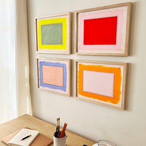 K&ouml;nnte beinhalten: Vier gerahmte Kunstdrucke mit farbenfrohen rechteckigen Designs in Rot, Gr&uuml;n, Orange und Blau. Das Kunstwerk wird an einer wei&szlig;en Wand ausgestellt. Die Rahmen sind aus hellem Holz gefertigt. Ein Schreibtisch mit Notizbuch, Stift und Kunstbedarf befindet sich im Vordergrund.
