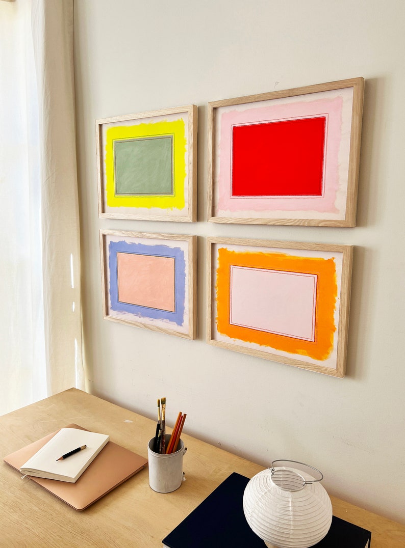 K&ouml;nnte beinhalten: Vier gerahmte Kunstdrucke mit farbenfrohen rechteckigen Designs in Rot, Gr&uuml;n, Orange und Blau. Das Kunstwerk wird an einer wei&szlig;en Wand ausgestellt. Die Rahmen sind aus hellem Holz gefertigt. Ein Schreibtisch mit Notizbuch, Stift und Kunstbedarf befindet sich im Vordergrund.