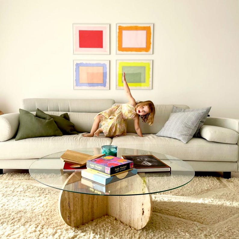 K&ouml;nnte beinhalten: Ein Wohnzimmer mit beigem Sofa, einem Glastisch und einem Shaggy-Teppich. Vier gerahmte abstrakte Kunstwerke h&auml;ngen an der Wand. Ein Kind ist auf dem Sofa. B&uuml;cher und eine Kiste mit Kunstbedarf liegen auf dem Tisch.