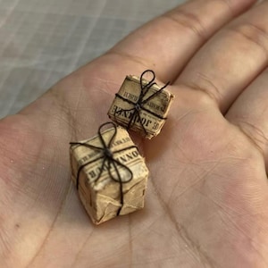 Può includere: Due pacchetti quadrati in miniatura avvolti in carta invecchiata color bianco sporco con testo nero stampato. Ogni piccolo pacco è legato con un filo nero sottile a forma di croce, creando un fiocco decorativo sulla parte superiore.
