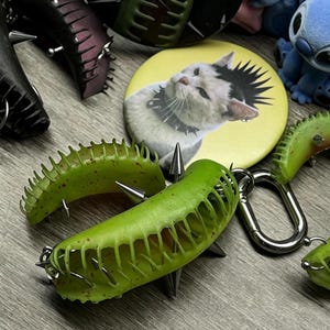 Può includere: Una collezione di portachiavi verdi a forma di dionaea con punte in metallo argentato, una spilla rotonda gialla raffigurante un gatto bianco con cresta nera e collare borchiato, e una statuetta blu visibile sullo sfondo dell'immagine.