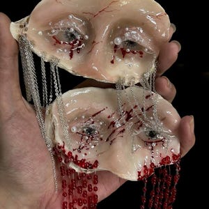 May include: Two pale, flesh-toned sculpted shells featuring realistic eyes with white pearl accents, red painted veins, and simulated blood drips. Silver chains and strands of small red beads hang from the edges of the shells, creating a layered effect.