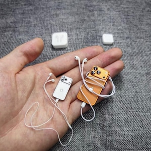 Può includere: Due repliche di smartphone in miniatura, una argentata e una arancione, ciascuna dotata di una fotocamera a tre lenti e un logo centrale. Entrambi i dispositivi sono collegati a auricolari bianchi con cavi lunghi. Due piccoli blocchi bianchi.