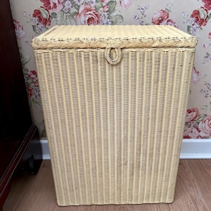 May include: A rectangular laundry hamper constructed from light beige woven wicker. The hamper features a flat, hinged lid with a braided border and a small, circular woven handle centred on the front edge for opening the lid with ease.