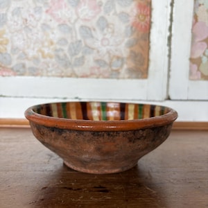 May include: A small, rustic earthenware bowl. The exterior is a warm brown, while the interior has a striped pattern of orange, green, and brown. The rim is a darker brown. The bowl is approximately 15 cm in diameter.