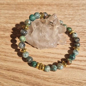May include: A beaded bracelet featuring round, speckled beads in shades of green, gray, and brown. Gold-colored spacers are interspersed between the beads. The bracelet is displayed on a wooden surface.