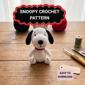 May include: A handmade crocheted Snoopy dog, white with black ears and nose, and a red collar, sits on a wooden surface. The image features the text "Snoopy Crochet Pattern" and "Easy to Download".
