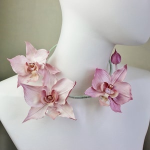 May include: A delicate pink orchid flower necklace with a pearl accent. The necklace is made of fabric flowers and is a beautiful and unique piece of jewelry.