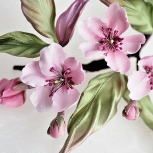 May include: Close-up of a floral arrangement featuring delicate pink blossoms with dark red centers and green leaves. The flowers are in various stages of bloom, with some buds still closed. The arrangement has a soft, romantic aesthetic.
