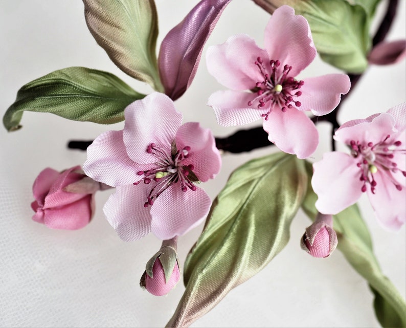 May include: Close-up of a floral arrangement featuring delicate pink blossoms with dark red centers and green leaves. The flowers are in various stages of bloom, with some buds still closed. The arrangement has a soft, romantic aesthetic.