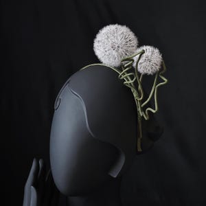 May include: A black headpiece featuring two large white dandelion seed heads and green wire stems. The headpiece is displayed on a black mannequin head. The dandelion seed heads are round and fluffy, with twisted wire stems.