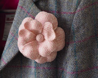 May include: A light pink fabric flower brooch with a textured herringbone pattern pinned to a grey wool blazer. The blazer features a subtle blue and pink plaid check pattern and a bright pink inner lining with a visible Harris Tweed label.