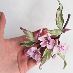 May include: A handcrafted floral branch with pink blossoms and green leaves. The delicate flowers have multiple petals and a dark pink center. The leaves have a subtle green and pink gradient. The stem is a dark brown color.
