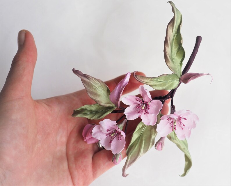 May include: A handcrafted floral branch with pink blossoms and green leaves. The delicate flowers have multiple petals and a dark pink center. The leaves have a subtle green and pink gradient. The stem is a dark brown color.