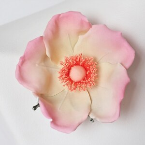 May include: A delicate, hand-crafted flower brooch with a pink and cream color gradient. The center of the flower features a bright orange, fluffy texture.