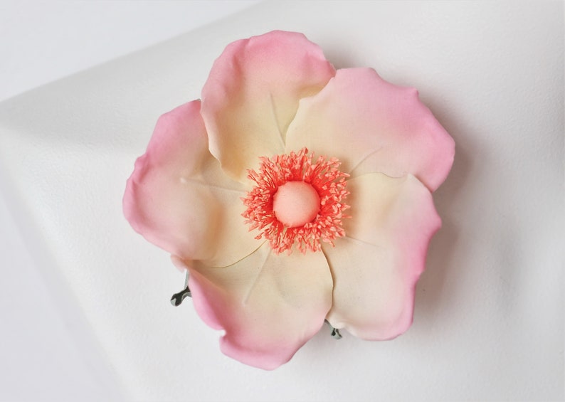 May include: A delicate, hand-crafted flower brooch with a pink and cream color gradient. The center of the flower features a bright orange, fluffy texture.