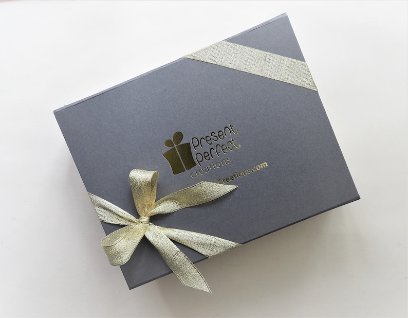 Op de afbeelding: Een grijze geschenkdoos met een gouden lint en strik. De doos heeft de tekst "Present Perfect Creations" en "creations.com" in goud. Het lint is diagonaal om de doos gewikkeld.