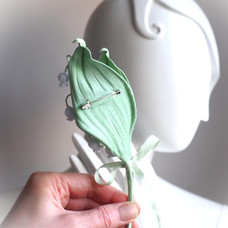 Op de afbeelding: Lichtgroene lelietje-van-dalen-broche met een zilveren speld. De broche heeft een groot, getextureerd blad met kleine witte bloemen en een groen lint. De broche wordt voor een witte mannequinhoofd gehouden.
