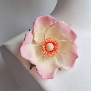 May include: A delicate, hand-crafted flower brooch with a pink and cream color gradient. The center of the flower is a fluffy, peach-colored pompom.