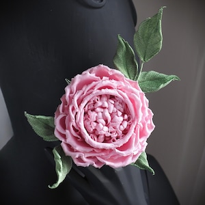 May include: A pink fabric rose with green fabric leaves. The rose is made of layers of fabric and has a ruffled appearance. The leaves are attached to the stem of the rose.