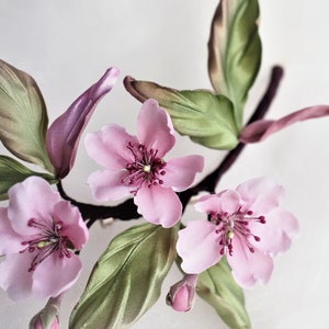 May include: Close-up of a floral arrangement featuring delicate pink blossoms with dark red centers and green leaves. The flowers are attached to a dark brown stem, with a few buds visible. The petals have a soft, silky texture.