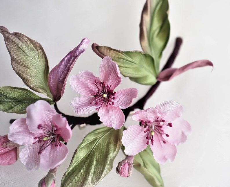 May include: Close-up of a floral arrangement featuring delicate pink blossoms with dark red centers and green leaves. The flowers are attached to a dark brown stem, with a few buds visible. The petals have a soft, silky texture.