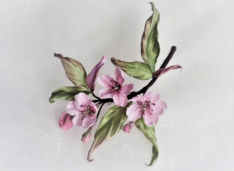 May include: A handcrafted floral arrangement featuring delicate pink blossoms, buds, and green leaves with brown accents. The artificial flowers are arranged on a dark brown stem, creating a natural and elegant aesthetic.