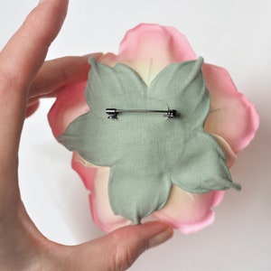 May include: A pink and white fabric flower with a green fabric backing and a metal pin on the back.